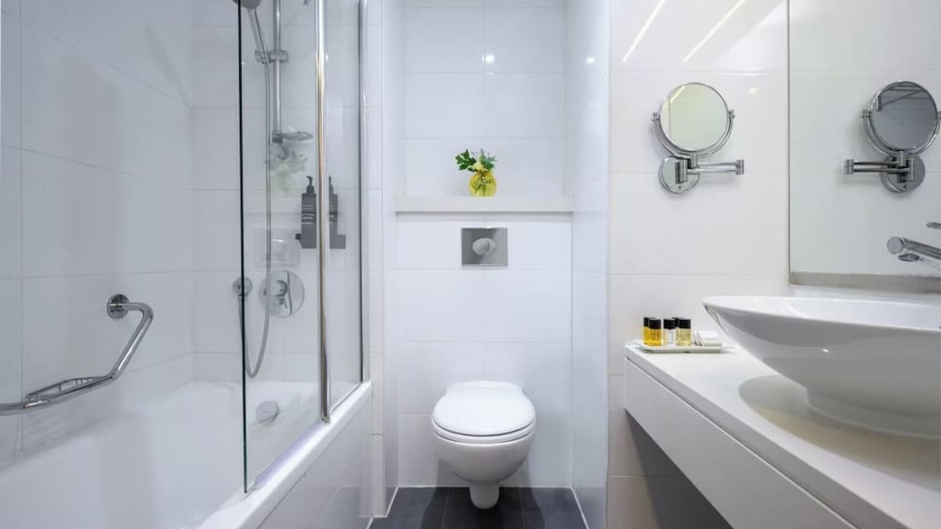 Modern bathroom with a bathtub and glass shower screen, wall-mounted toilet, white vanity basin, magnifying mirrors, and toiletries at Prima Park Hotel.