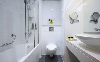 Modern bathroom with a bathtub and glass shower screen, wall-mounted toilet, white vanity basin, magnifying mirrors, and toiletries at Prima Park Hotel.