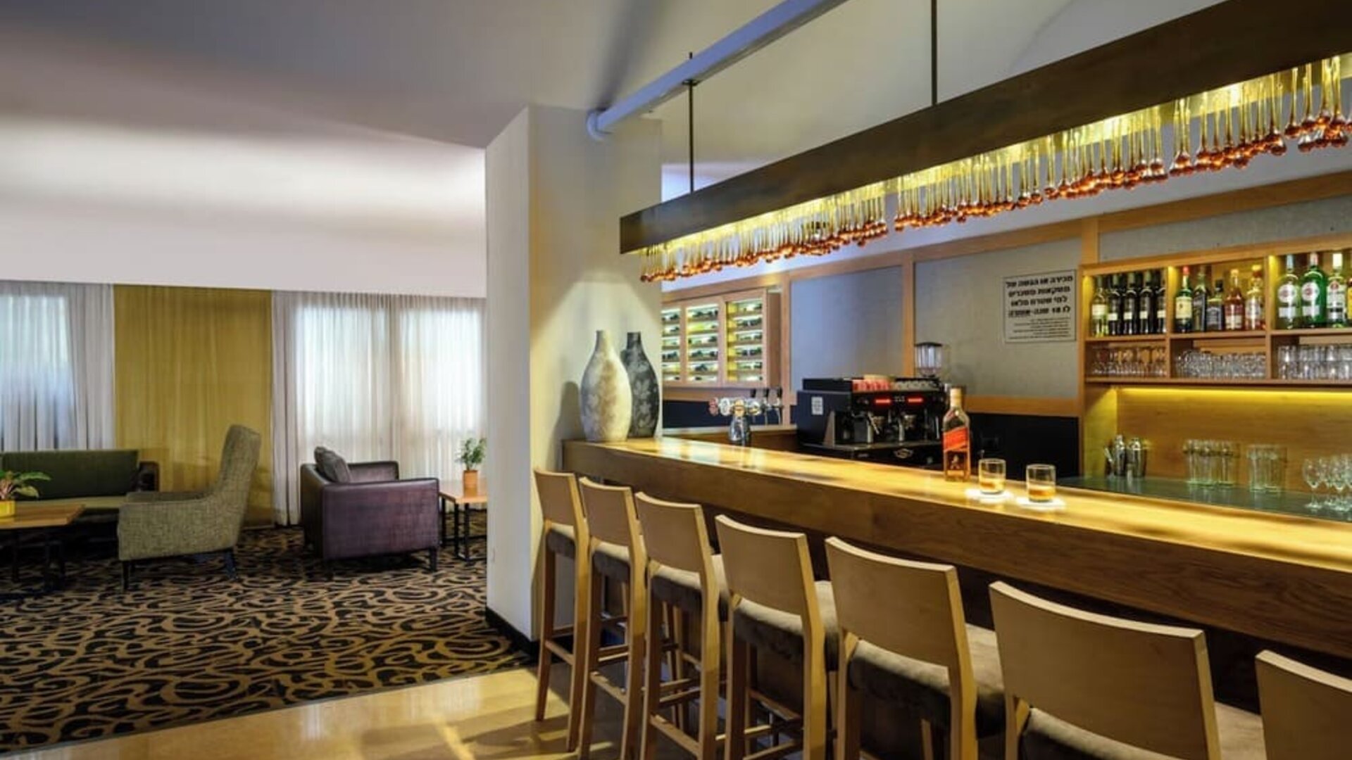 Hotel bar with a long wooden counter, bar stools, illuminated bottle shelves, glassware, and an adjoining lounge seating area at Prima Park Hotel.