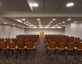 Meeting room arranged classroom-style with rows of chairs, patterned carpeting, a presentation area, and overhead lighting at Prima Park Hotel.