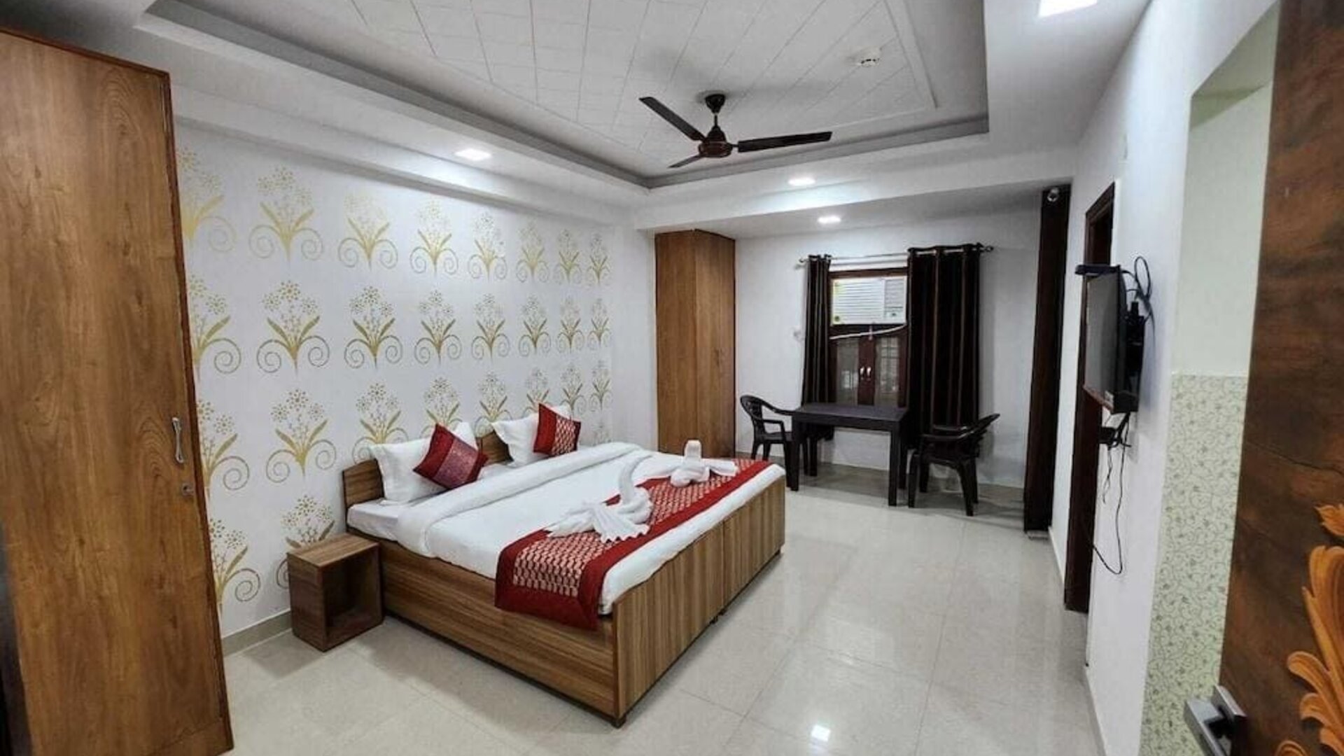 Guest room with a double bed, white bedding, brown and gold accent cushions, a matching bed runner, wooden wardrobe, patterned floral feature wall, and a small seating area by the window at Hotel Urban Abode.