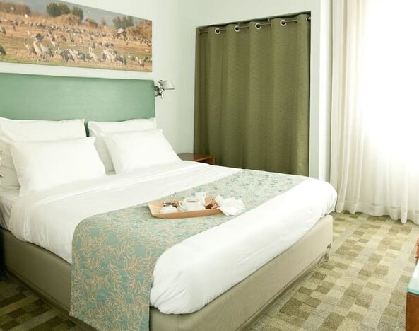 Guest bedroom featuring a king bed, sage-green headboard, patterned runner and bright windows at Prima Galil Tiberias.