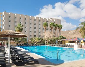 Outdoor swimming pool with rows of sun loungers, parasols and views of the hotel building and surrounding desert hills at Prima Music Eilat.