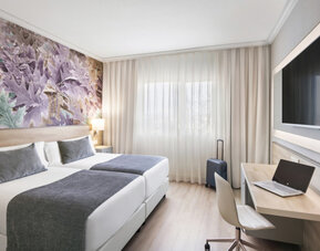 Guest room with twin beds, grey bedding, a work desk, open suitcase and decorative botanical wall art at Hotel Sallés Ciutat del Prat.