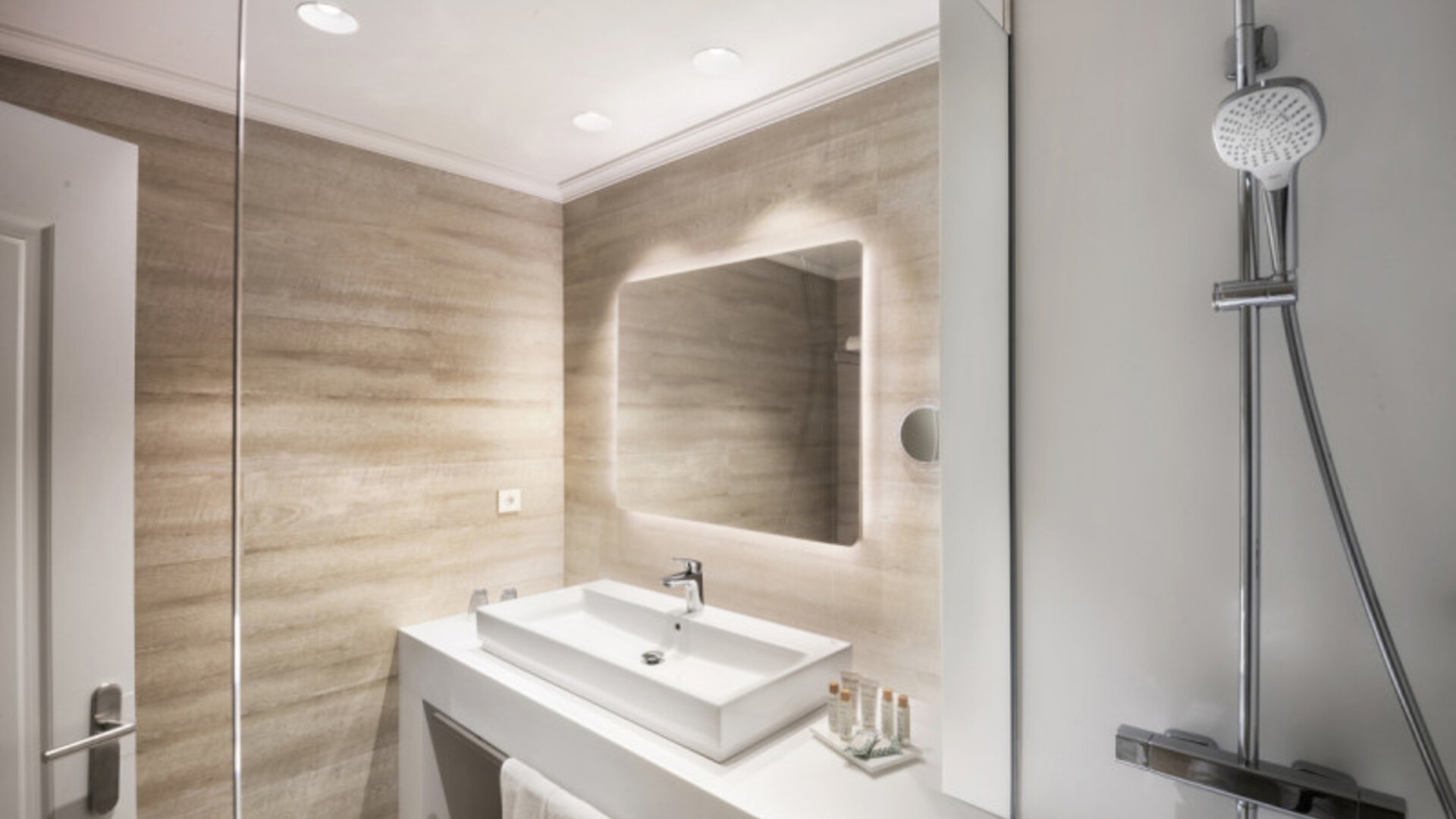 Modern bathroom with a glass shower enclosure, white vanity basin, illuminated mirror and beige tiled walls at Hotel Sallés Ciutat del Prat.