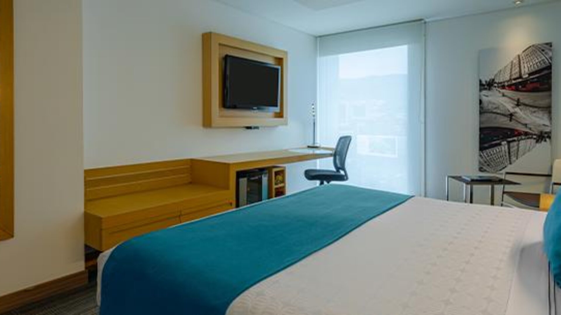 Guest room with white bedding, teal accent cushions, wooden desk and large window at Sonesta Hotel Bogotá.