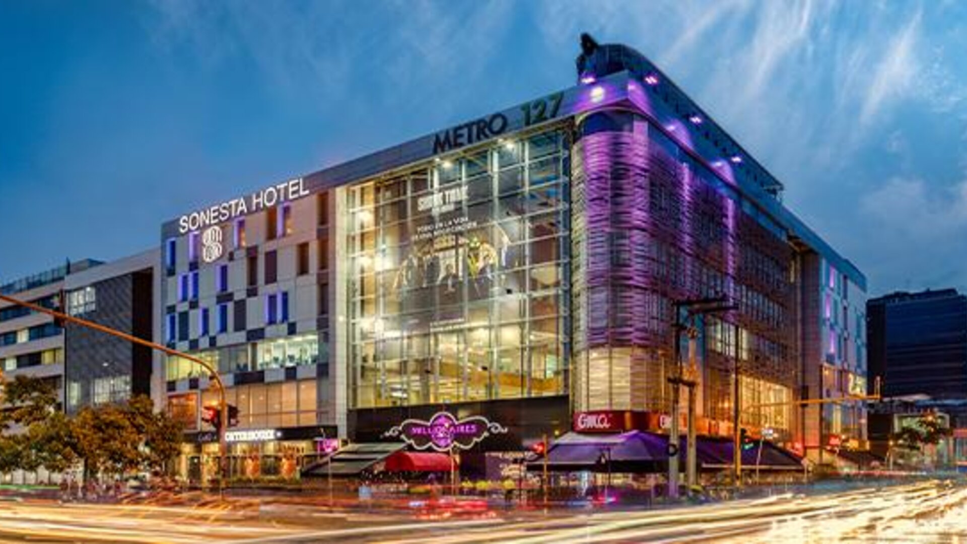 Modern glass-fronted hotel exterior illuminated with purple lighting against the evening sky at Sonesta Hotel Bogotá.