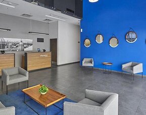 Hotel reception with wood-fronted desks, grey sofas, round wall mirrors and a bright blue accent wall at City Express by Marriott CDMX La Villa.