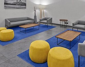 Lounge with grey sofas, yellow round ottomans, wooden coffee tables and blue carpeting at City Express by Marriott CDMX La Villa.