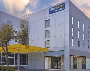 Modern hotel exterior with silver panelled walls, large windows and a yellow entrance canopy at City Express by Marriott CDMX La Villa.