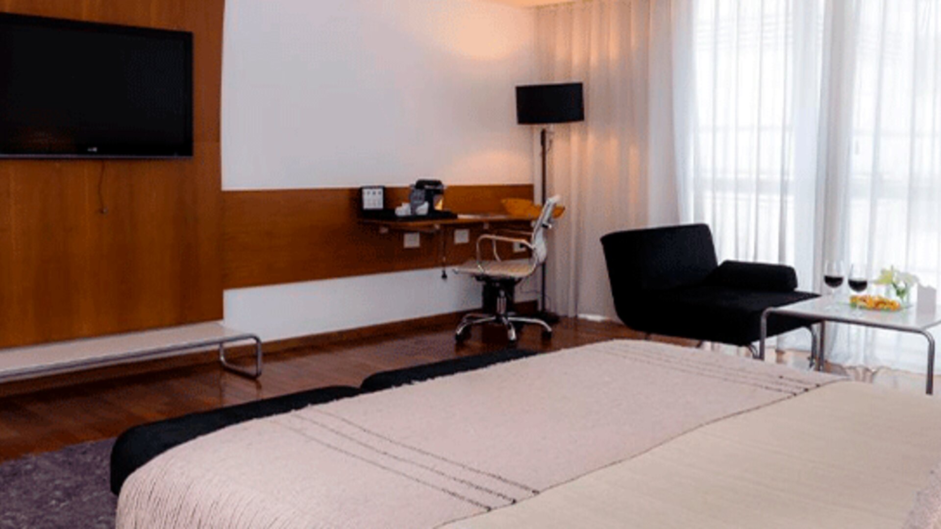 Guest room with large bed, wall-mounted television, desk, black lounge chairs and floor-to-ceiling curtains at Design Suites Buenos Aires.