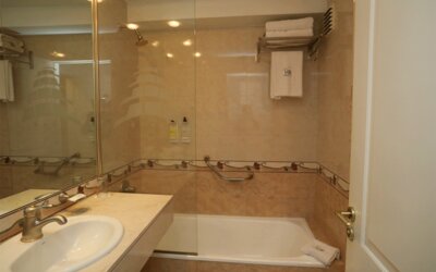 Bathroom with beige tiled walls, glass shower screen, bathtub, white vanity sink and grab rails at HR Esmeralda Luxor.