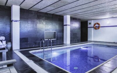Indoor swimming pool with blue tiled water, dark stone walls, metal handrails and a towel rack at Hotel Best Western PLUS Alfa Aeropuerto.