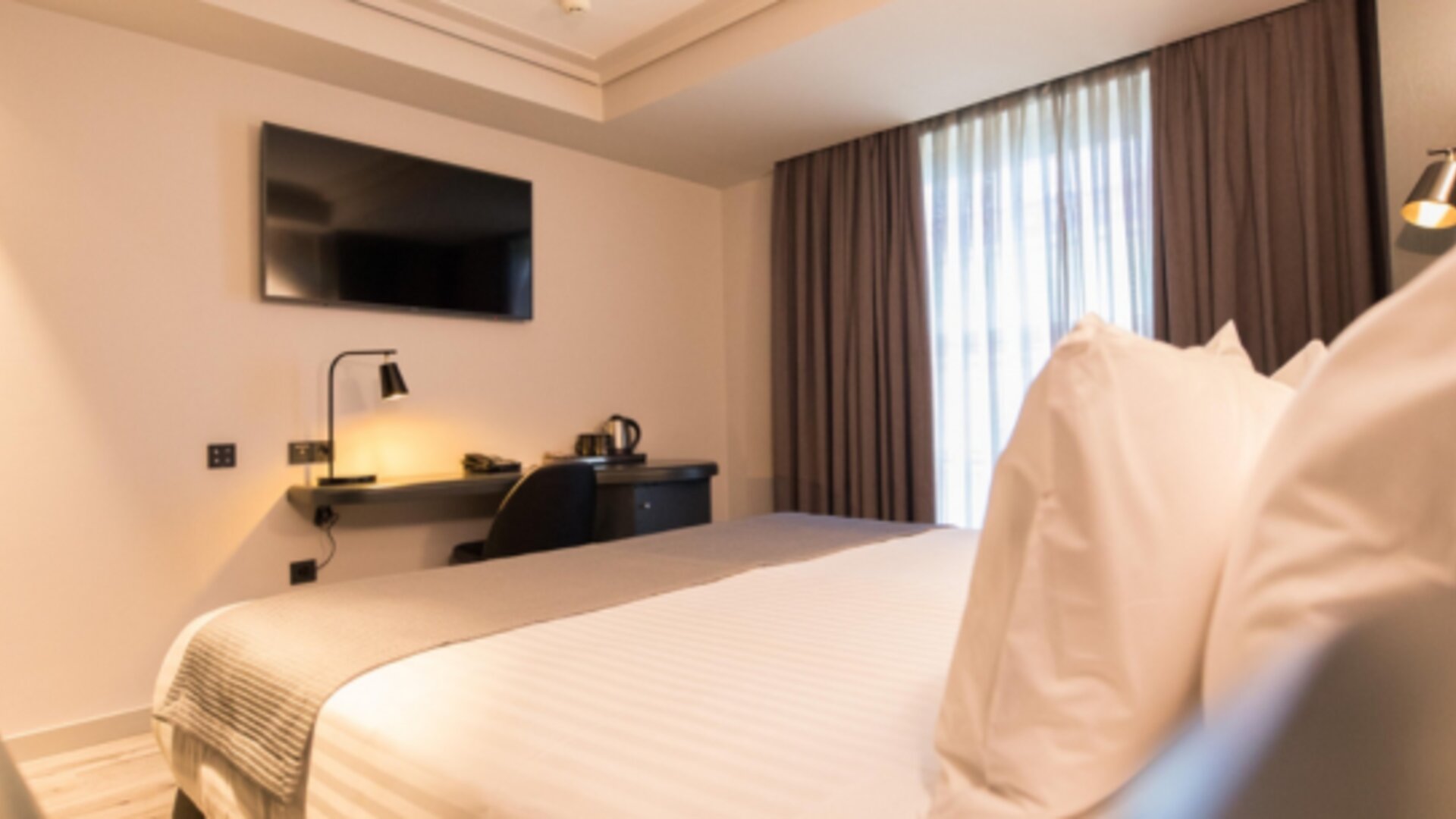Guest room with white bedding, wall-mounted television, desk, dark curtains and bedside lighting at Hotel Silken Saaj Las Palmas.