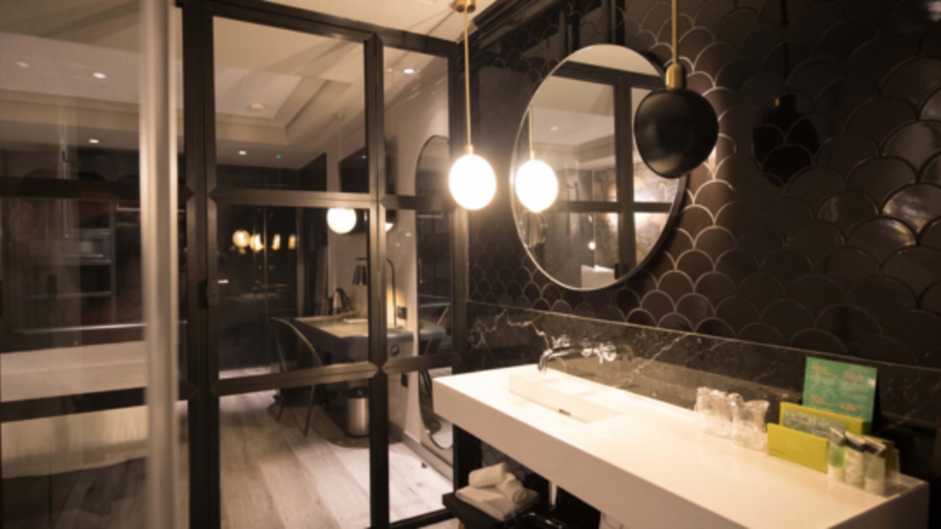 Bathroom vanity with white countertop, round mirror, dark scalloped wall tiles and glass shower enclosure at Hotel Silken Saaj Las Palmas.