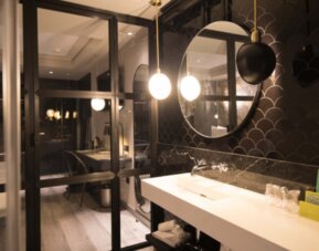 Bathroom vanity with white countertop, round mirror, dark scalloped wall tiles and glass shower enclosure at Hotel Silken Saaj Las Palmas.