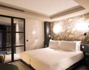 Guest room with white bed, patterned feature wall, bedside lighting and black-framed glass doors at Hotel Silken Saaj Las Palmas.