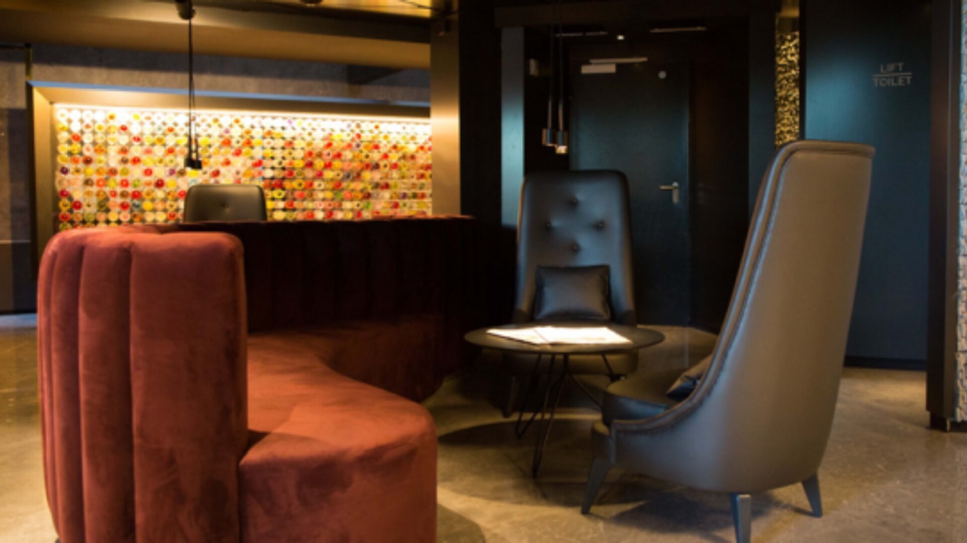 Hotel lounge with curved burgundy sofa, grey armchairs, round coffee table and illuminated decorative wall at Hotel Silken Saaj Las Palmas.