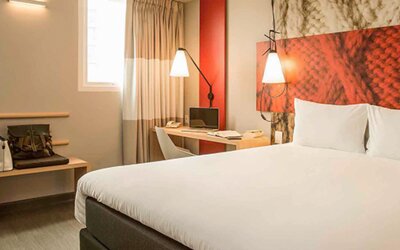 Guest room with a large white bed, red accent headboard, bedside lamps, desk and curtained windows at Ibis Lima Reducto Miraflores.