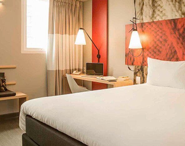 Guest room with a large white bed, red accent headboard, bedside lamps, desk and curtained windows at Ibis Lima Reducto Miraflores.
