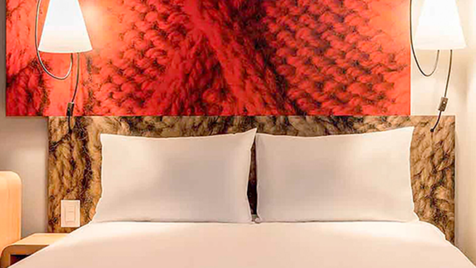 Close view of a double bed with white pillows, red decorative headboard and matching wall lamps at Ibis Lima Reducto Miraflores.