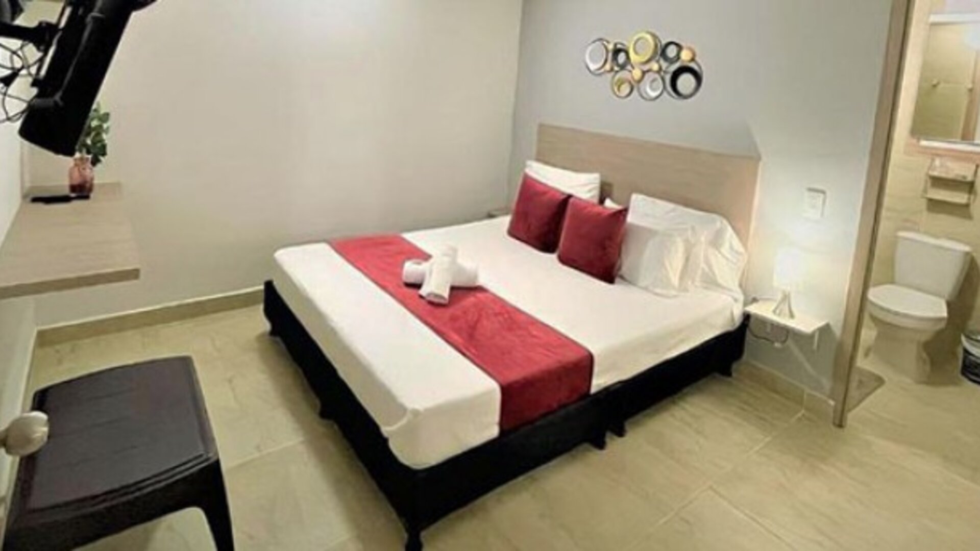 Guest room with double bed, red runner and cushions, wall-mounted television and ensuite bathroom visible through the doorway at 12 Nights Hotel.