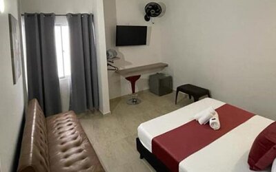 Guest room with double bed, red runner and cushions, brown sofa, wall-mounted television and curtained window at 12 Nights Hotel.