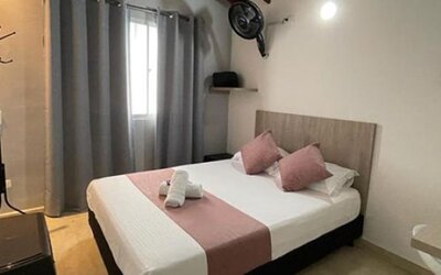Guest room with double bed, pink accent cushions and runner, grey curtains, wall fan and wooden desk at 12 Nights Hotel.
