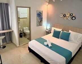 Guest room with double bed, teal runner and cushions, bedside lighting, desk and ensuite bathroom at 12 Nights Hotel.