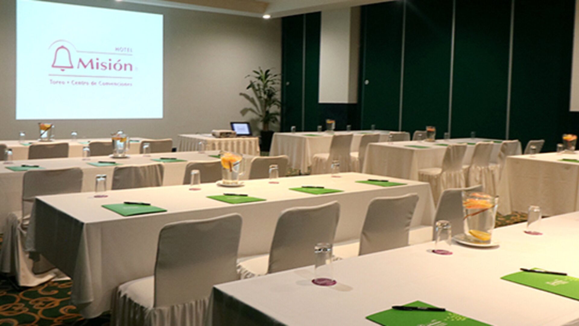 Conference room with U-shaped tables, white tablecloths, green notepads and a presentation screen at Hotel Misión Toreo Centro De Convenciones.