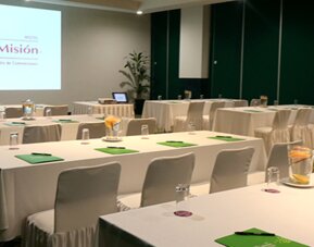 Conference room with U-shaped tables, white tablecloths, green notepads and a presentation screen at Hotel Misión Toreo Centro De Convenciones.
