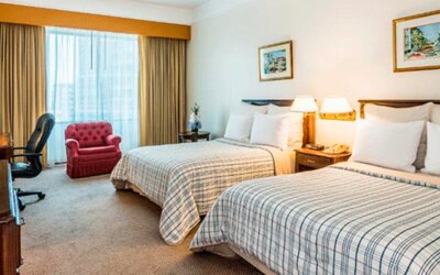 Twin guest room with two beds, striped bedding, red armchair, desk, television and large curtained window at Hospedium Princess Hotel Panamá.