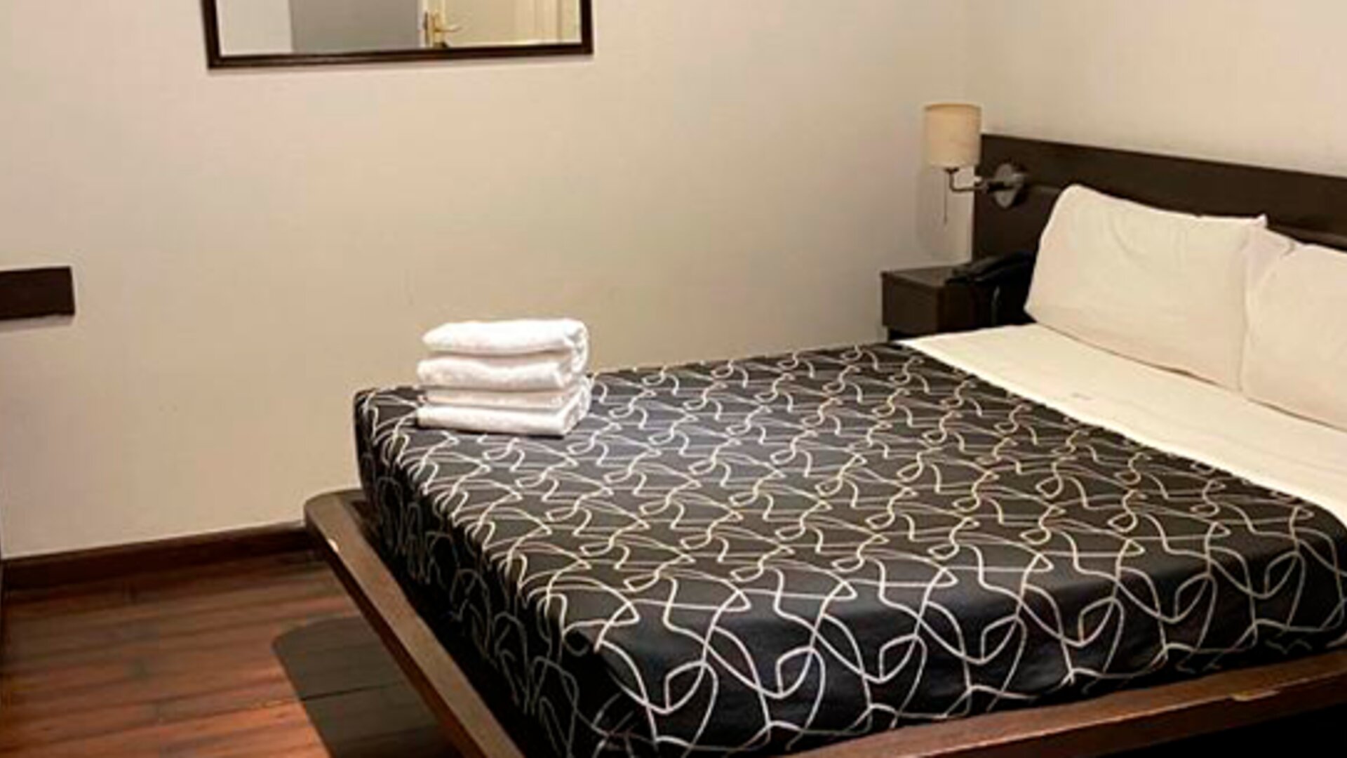 Guest room with double bed, black patterned bedding, folded white towels, bedside table and wall mirror at Bagu Buenos Aires.