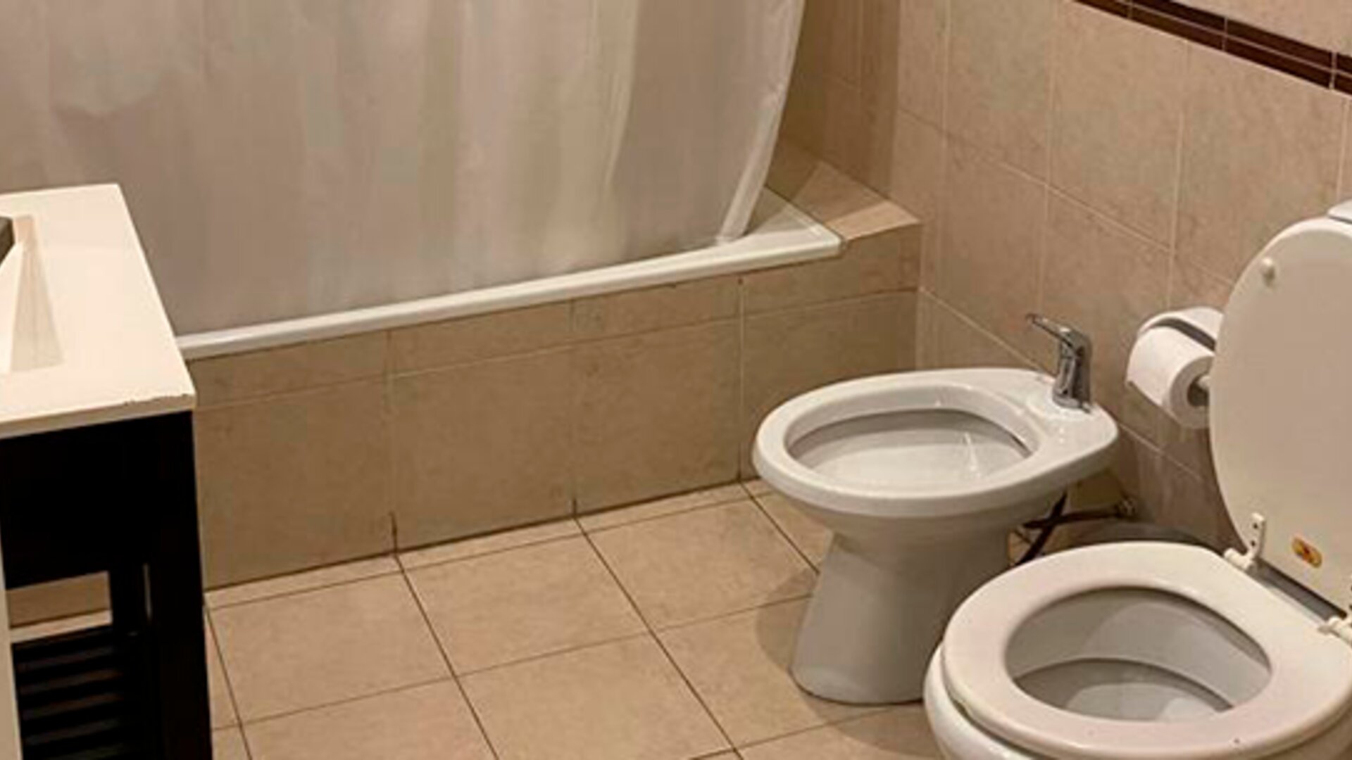 Bathroom with bathtub and shower curtain, toilet, bidet and beige tiled walls and flooring at Bagu Buenos Aires.