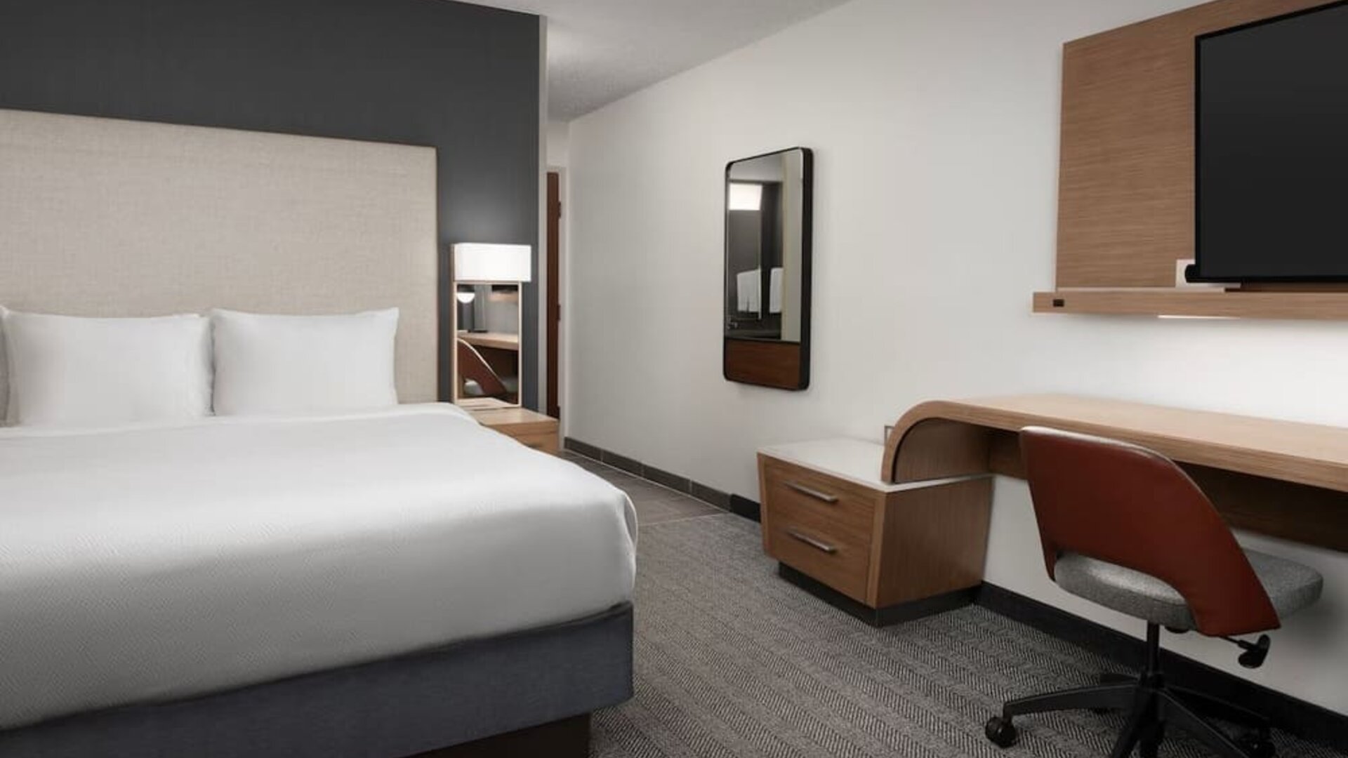 Guest room with a white bed, upholstered headboard, wooden desk, office chair, wall-mounted television and gray carpeting at Courtyard By Marriott Dothan.