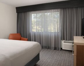 Guest room with white bed, orange accent chair, large curtained window, television and wooden desk at Courtyard By Marriott Dothan.