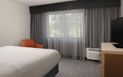 Guest room with white bed, orange accent chair, large curtained window, television and wooden desk at Courtyard By Marriott Dothan.
