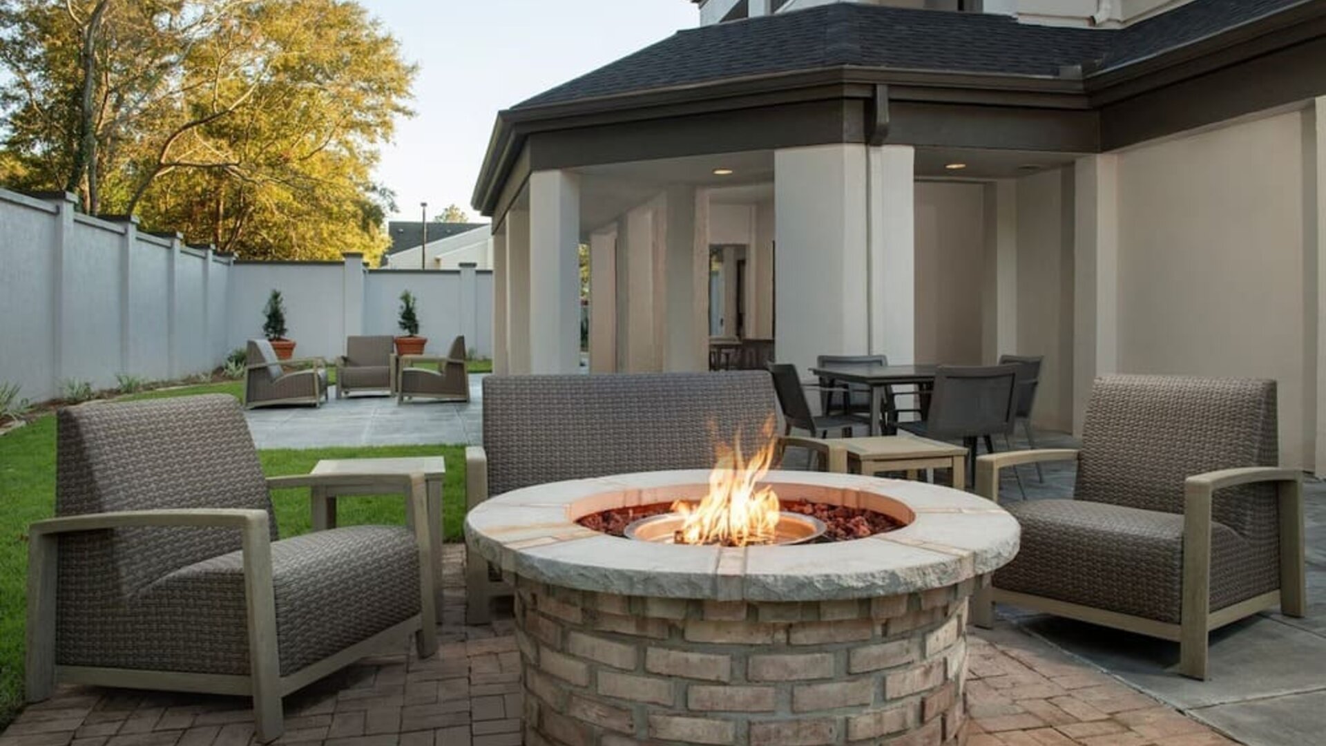 Outdoor patio with cushioned lounge chairs surrounding a circular brick fire pit, with lawn and landscaping at Courtyard By Marriott Dothan.