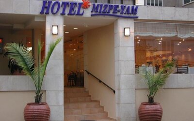 Hotel entrance with stone facade, glass doors, exterior signage and large potted palm plants at Mizpe Yam Netanya Boutique Hotel.