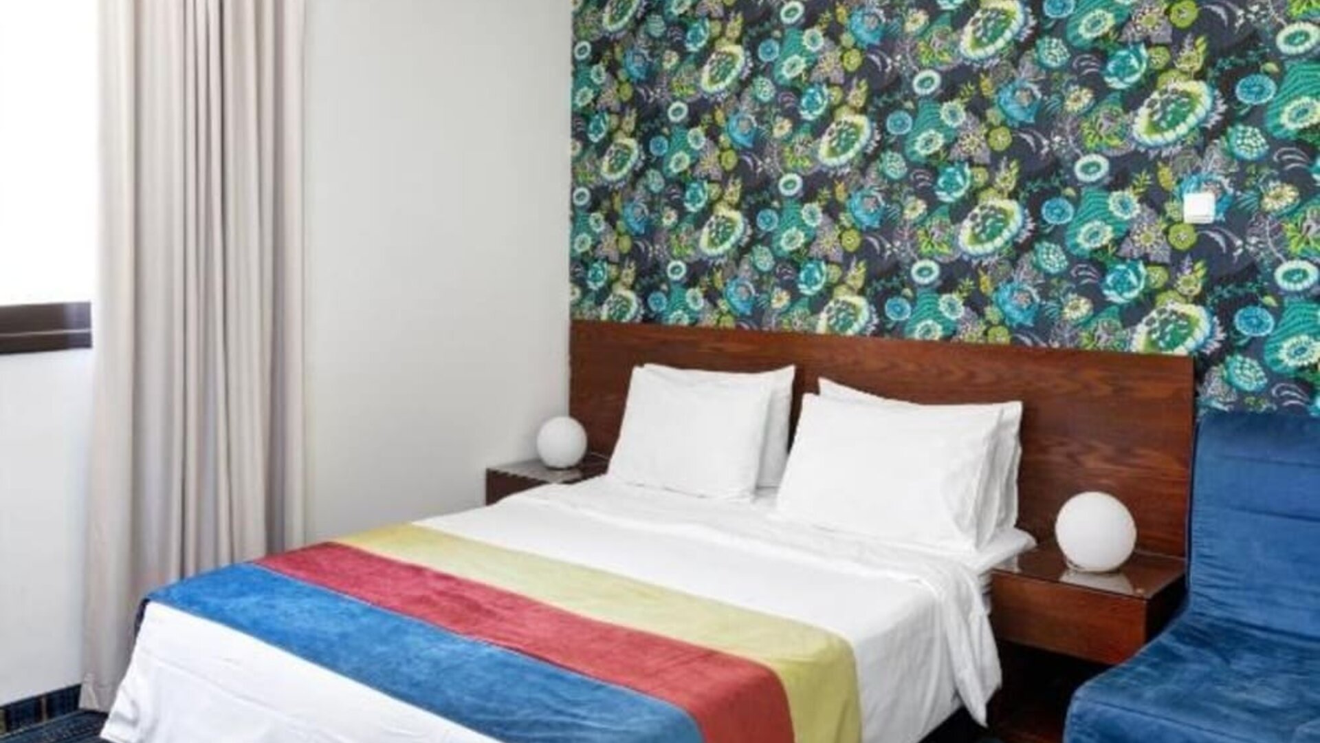 Guest room with double bed, colourful patterned feature wall, blue armchair and striped carpet at Mizpe Yam Netanya Boutique Hotel.