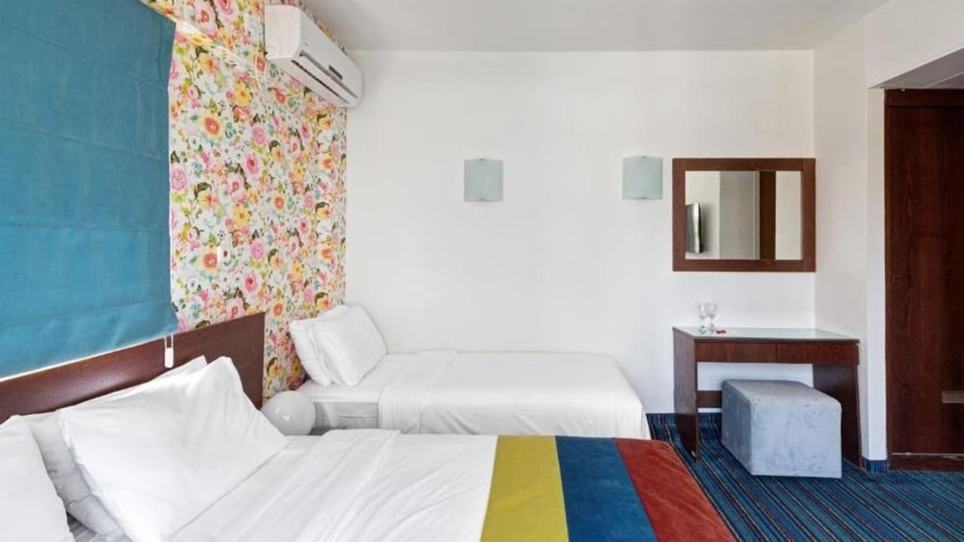 Guest room with two beds, colourful floral feature wall, blue curtains and matching striped carpet at Mizpe Yam Netanya Boutique Hotel.
