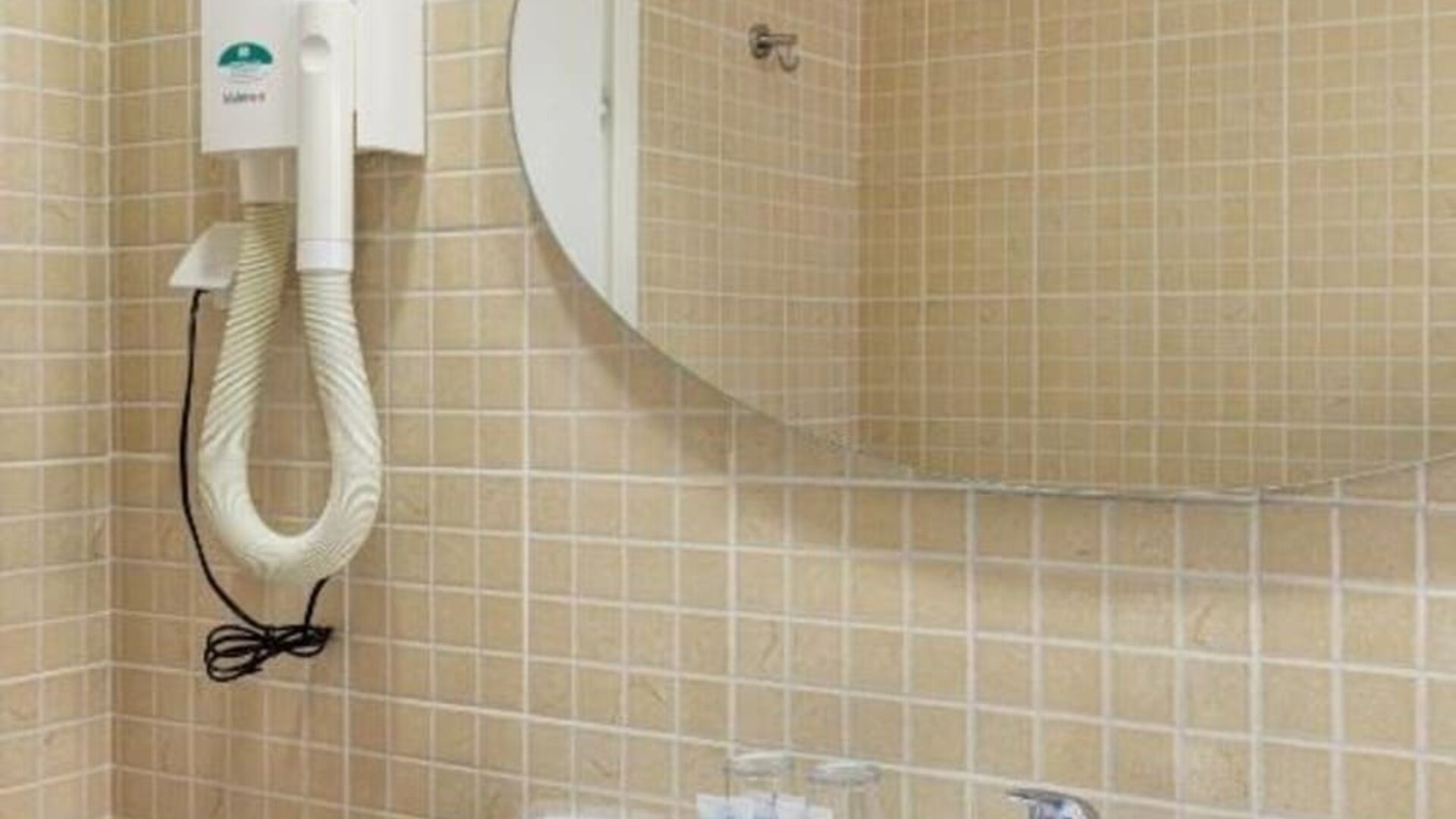 Bathroom with beige tiled walls, white sink, oval mirror and wall-mounted hairdryer at Mizpe Yam Netanya Boutique Hotel.