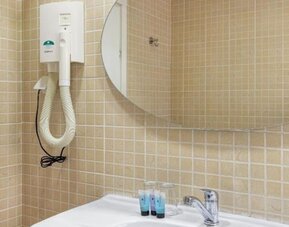 Bathroom with beige tiled walls, white sink, oval mirror and wall-mounted hairdryer at Mizpe Yam Netanya Boutique Hotel.