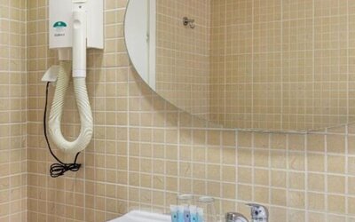 Bathroom with beige tiled walls, white sink, oval mirror and wall-mounted hairdryer at Mizpe Yam Netanya Boutique Hotel.