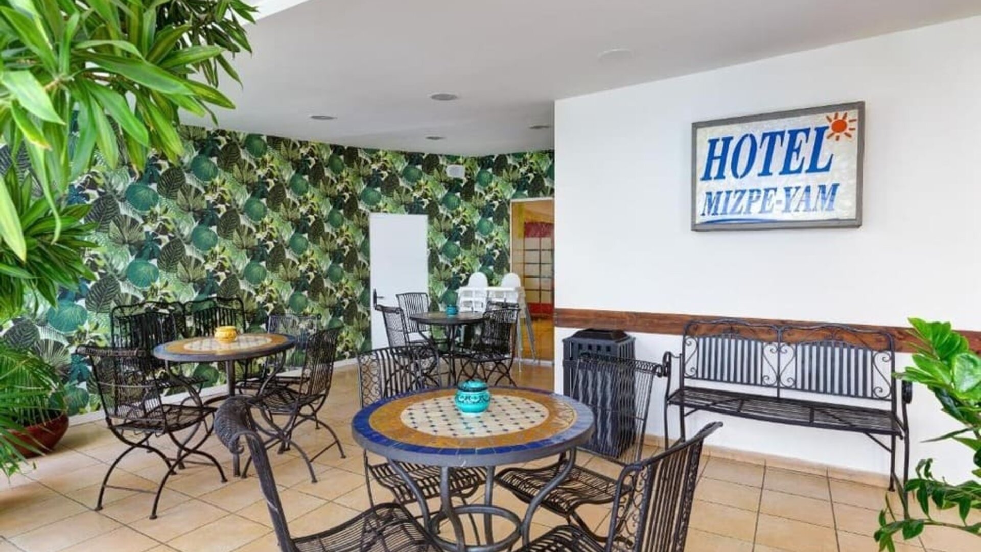 Hotel seating area with round mosaic tables, wrought-iron chairs, leafy wallpaper and a wall sign at Mizpe Yam Netanya Boutique Hotel.