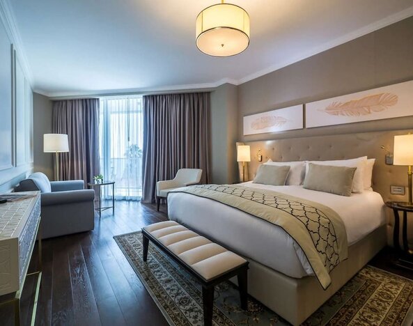 Guest room with king bed, upholstered headboard, bedside lamps, seating area and large curtained windows at David Tower Hotel Netanya.