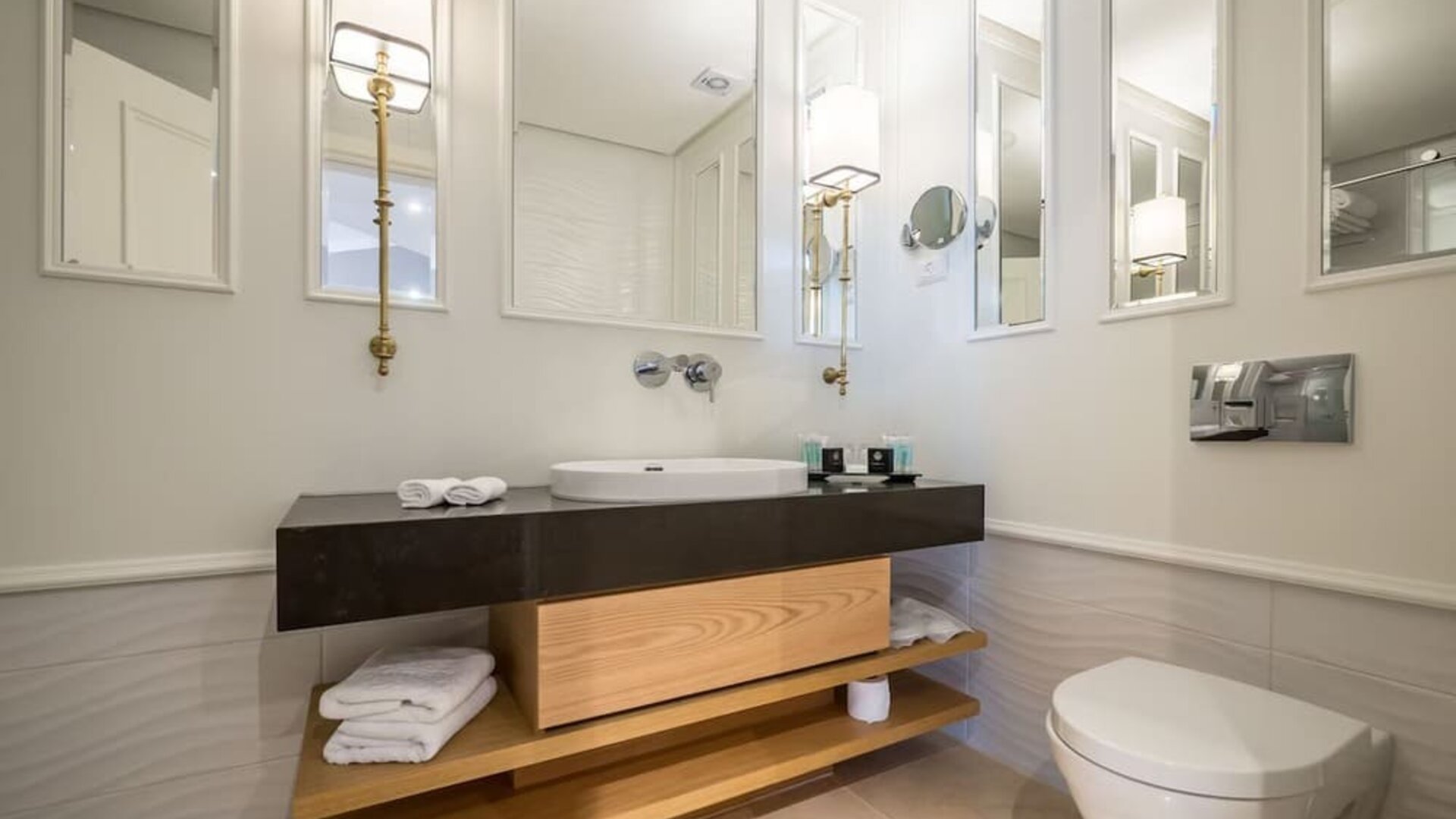 Modern bathroom with dark vanity, white basin, illuminated mirrors, folded towels and wall-mounted toilet at David Tower Hotel Netanya.