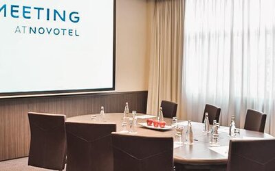 Meeting room with a conference table, brown chairs, presentation screen and large windows with sheer curtains at Novotel Lima San Isidro.