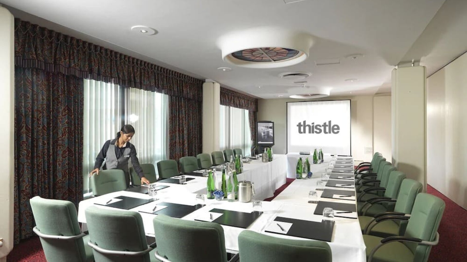 Room for business meetings with large windows, screen, and seating around single table for around a dozen people.