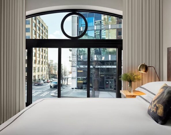 Bedroom with large window showing city street view, white bedding, and neutral decor at Kimpton Palladian Hotel.